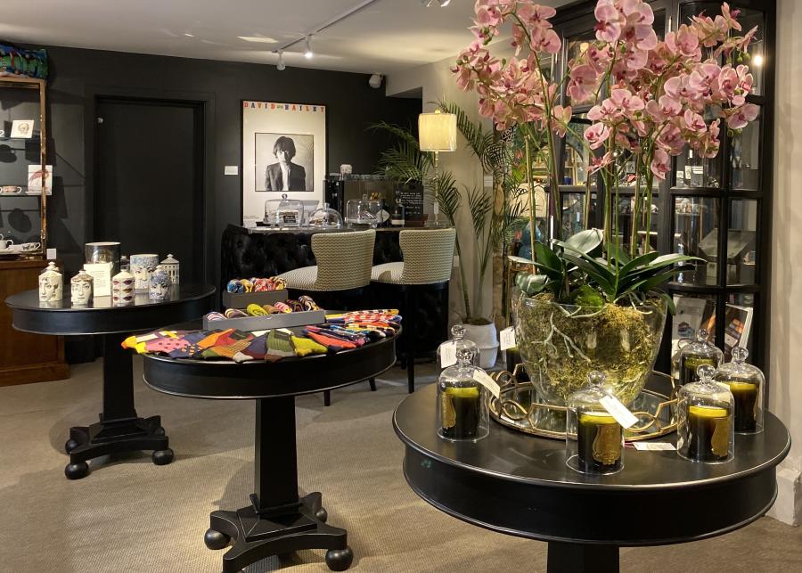 Cotswold Stay - Wonderful Winchcombe Interior of Barnbury, a stylish independent shop in Winchcombe. In the foreground, a large glass planter filled with pink orchids sits on a round black display table surrounded by decorative candles. Nearby tables display colorful patterned socks and home interior items. In the back, two upholstered chairs sit at a small counter beneath a black-and-white portrait poster, with shelves of home décor and plants throughout the space.