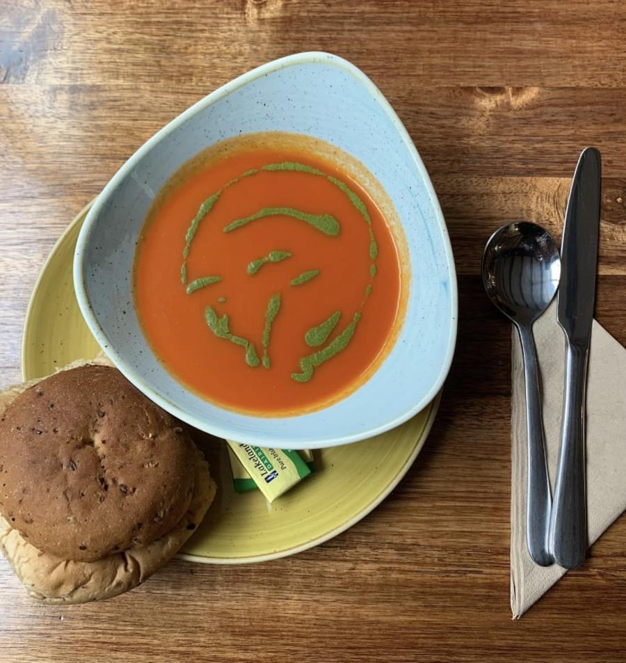 Cotswold Stay - Wonderful Winchcombe Overhead view of a place setting on a wooden table featuring a bowl of smooth tomato soup drizzled with green herb oil. The bowl sits on a light green plate, accompanied by a bread roll. To the right are a neatly placed knife and fork on a folded napkin, along with a small condiment container. The scene is warmly lit, highlighting the textures of the wood and the vibrant color of the soup.