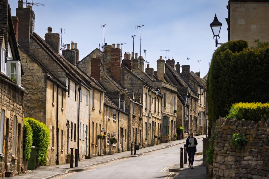 Cotswold Stay - Wonderful Winchcombe A narrow, gently curving street lined with closely packed stone houses featuring sloped roofs, chimney stacks, and small windows. The buildings vary in warm neutral tones like beige, cream, and light brown, with some greenery climbing walls and hedges along the edges. A single pedestrian walks along the pavement on the right side of the road. A low stone wall and neatly trimmed bush appear in the foreground, while traditional street lamps rise above the homes. The sky overhead is pale and clear, casting soft daylight across the quiet scene in Winchcombe.