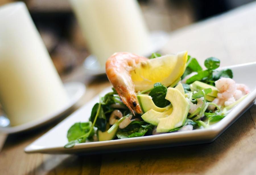 Cotswold Stay - Wonderful Winchcombe White rectangular plate holding a composed salad with sliced avocado, leafy greens, and pieces of seafood, accompanied by a lemon wedge. The dish is placed on a wooden table, with a softly blurred background featuring a white candle and dining setting, creating a warm restaurant atmosphere.