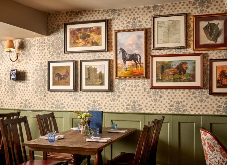 Cotswold Stay - Wonderful Winchcombe Inside the cosy restaurant at The Lion Inn. A wooden table set for four, surrounded by dark wooden chairs. The wall behind features patterned wallpaper and is decorated with a gallery of framed artwork, many depicting horses and animals in various styles and sizes. A small vase of yellow flowers sits on the table alongside menus, plates, and cutlery. Warm, ambient lighting creates an inviting, traditional atmosphere.